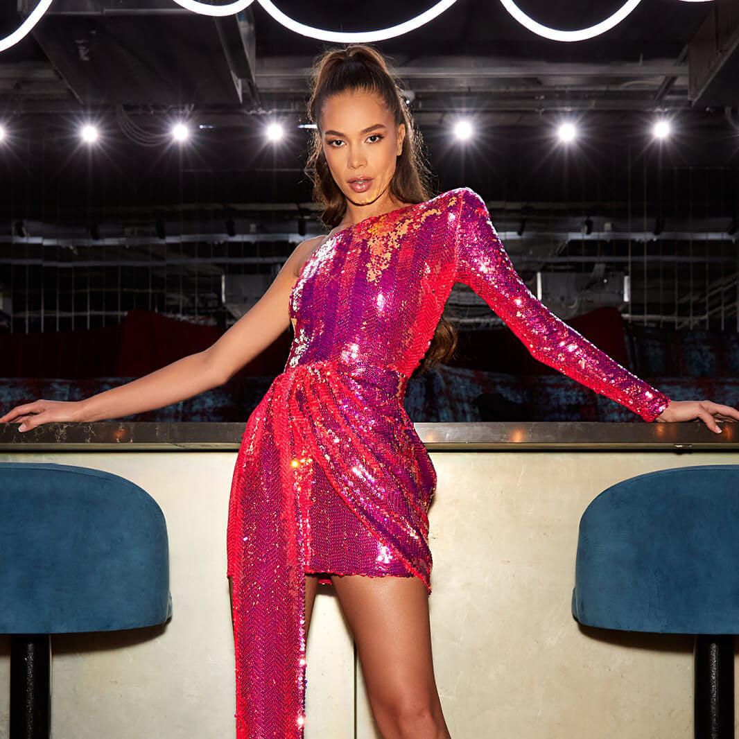 Model in pink one-shoulder sequin dress posing at a bar with blue chairs.